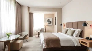 Modern hotel room with comfortable bed, neutral tones, natural lighting from large window, contemporary furniture and clean minimalist decor, no visible text or signage