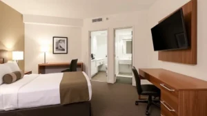 Modern hotel room with comfortable queen bed, work desk, flat-screen TV, and contemporary bathroom visible through open door. Neutral tones, clean and well-lit space with professional furnishings, no text or identifying marks visible.