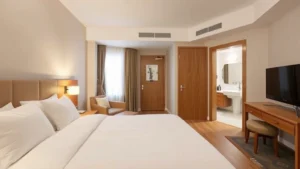 Modern hotel room interior with comfortable bed, clean white bedding, warm lighting, wooden furniture, and contemporary bathroom visible through doorway, bright and inviting residential space