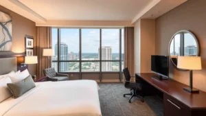 Modern hotel room interior with contemporary king bed, neutral beige and gray tones, sleek wooden desk workspace, floor-to-ceiling windows overlooking Orlando landscape, professional business traveler environment, soft warm lighting