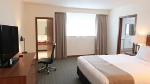 Bright, clean modern hotel room with queen bed, work desk, flat-screen TV, and contemporary bathroom visible through open door, neutral color palette, natural window light