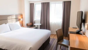 Modern budget hotel room with clean white bedding, functional furniture, and welcoming neutral décor. Natural window light illuminates comfortable sleeping space with digital alarm clock and simple desk area. Bright and inviting atmosphere showing well-maintained accommodation.