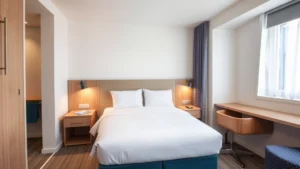 Modern budget hotel room with clean queen bed, bright white linens, functional nightstands, and contemporary lighting. Compact but well-organized layout showing efficient use of space. Neutral color palette with blue accents. Photorealistic, daytime interior shot.