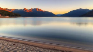 Serene waterfront view of Lake Chelan with sandy beach, calm blue water, and surrounding pine-covered mountains during golden hour sunset, no people visible, photorealistic landscape