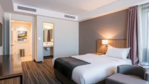 Modern hotel room interior with neatly made queen bed, contemporary furnishings, clean bathroom visible through open door, warm ambient lighting, no visible signage or numbers