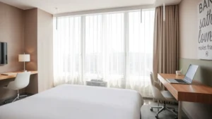 Modern hotel room with clean white bedding, natural sunlight streaming through large windows, desk workspace with laptop visible, contemporary minimalist decor, empty and ready for guests