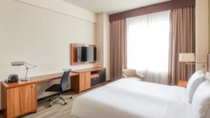 Modern hotel room interior with queen bed, flat-screen TV, work desk, and neutral beige color palette, bright natural light from window