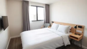 Modern economy hotel room with clean white bedding, comfortable mattress, simple furnishings, bright natural light from window, contemporary design without clutter, residential-quality comfort