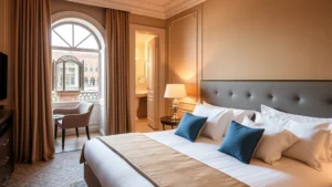 Modern hotel bedroom in Venice with queen bed, soft gold and blue Venetian color scheme, arched window overlooking canal water, contemporary bathroom visible through doorway, warm lighting from bedside lamps, luxurious linens and pillows, no visible text or signage