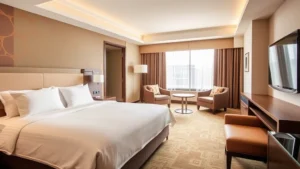 Modern hotel bedroom with premium bedding, soft lighting, and contemporary furnishings in warm earth tones, showing comfortable seating area and large window with natural light
