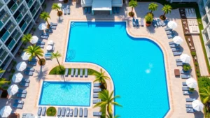 Aerial view of a modern hotel resort with sparkling blue Olympic-length swimming pool surrounded by lounge chairs, umbrellas, and palm trees on a sunny day