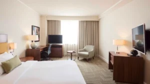 Spacious hotel bedroom with comfortable bed, desk workspace, modern furnishings, and natural window light in neutral tones