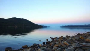Serene Lake Superior waterfront at sunset with calm waters, rocky shoreline, and forested hills reflected in glassy surface, golden hour lighting