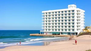 Bright, modern oceanfront hotel building with white and blue exterior, multiple stories with balconies overlooking Atlantic Ocean, sandy beach in foreground, clear blue sky, waves visible, beachgoers enjoying sand