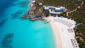 Aerial view of a pristine turquoise Mediterranean beach with white sand, crystal-clear shallow waters, and a luxury resort with white buildings nestled among green vegetation overlooking the coastline