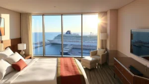 Modern hotel bedroom with ocean view overlooking cruise ship port, morning sunlight, contemporary furnishings, port visible through large windows
