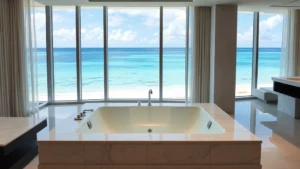 Modern luxury hotel suite with large sunken jacuzzi positioned near floor-to-ceiling windows overlooking tropical ocean coastline, contemporary bathroom with marble finishes and soft ambient lighting, empty and pristine