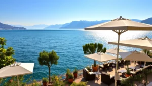 Serene lakefront hotel terrace with white umbrellas overlooking sparkling blue Lake Garda waters, Mediterranean gardens with lemon trees, comfortable lounge seating, golden hour sunlight reflecting on water, Italian Alps visible in distant background