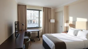 Modern hotel room interior with contemporary furnishings, clean lines, comfortable bed with quality linens, work desk, flat-screen TV, and neutral color palette, softly lit with natural window light showing urban Milan street view