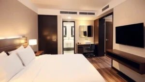 Modern hotel room with contemporary furnishings, comfortable bed with white linens, flat-screen TV mounted on wall, warm lighting, clean bathroom visible through open door, professional business workspace with desk and chair