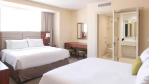Bright, modern hotel guest room with king bed, contemporary furnishings, and clean bathroom visible through open door. Natural light from window. Neutral color palette with warm tones. No visible text or signage.