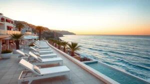 Beachfront Mediterranean hotel terrace with white loungers overlooking turquoise sea at sunset, palm trees visible, coastal cliff backdrop, golden hour lighting, no people visible, serene atmosphere