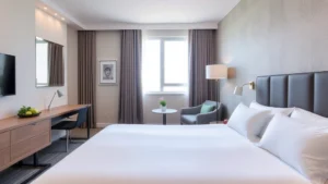 Modern hotel room with neatly made bed, comfortable pillows, neutral tones, clean windows showing daytime, contemporary furniture, welcoming atmosphere