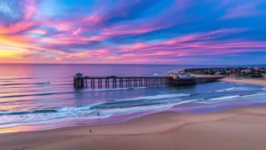 Aerial sunset view of Huntington Beach pier extending into the Pacific Ocean with surfers in the water, sandy beach in foreground, colorful sky reflecting on ocean, residential buildings visible inland