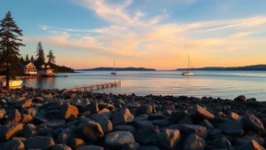 Serene waterfront view of Puget Sound with coastal homes and sailboats, rocky beach in foreground, pine trees, golden hour lighting, calm water reflecting sky