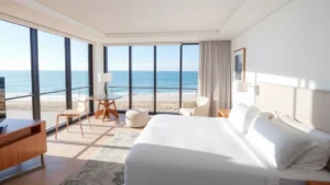 Modern oceanfront hotel room with contemporary furnishings, large windows overlooking calm blue ocean water, clean white bedding, minimalist design aesthetic, bright natural lighting from floor-to-ceiling windows, coastal beachfront view at daytime