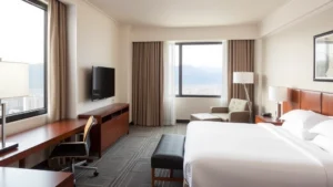 Modern hotel guest room with king bed, contemporary furnishings, neutral tones, clean lines, work desk, flat-screen TV mounted on wall, natural lighting from window showing mountain landscape outside