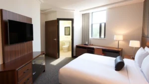 Modern hotel room with neatly made bed, work desk with lamp, flat-screen TV, and clean bathroom visible through open door, morning natural light streaming through window