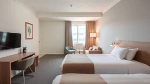 Modern budget hotel room with clean bed, neutral décor, bright natural light from window, comfortable seating area, contemporary furnishings, welcoming residential atmosphere, no visible text or signage