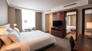 Modern hotel room with contemporary furnishings, king bed with white linens, work desk, flat-screen TV, and updated bathroom visible through open door, natural daylight from window