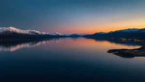 Panoramic view of Kachemak Bay at sunset with snow-capped mountains reflected in calm water, coastal village lights visible in distance, golden hour lighting, photorealistic landscape