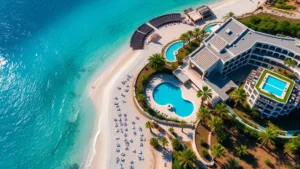Aerial view of a Mediterranean beachfront hotel with turquoise water, white sandy beach, and palm trees, showing terraced architecture and pool areas overlooking the Ionian Sea