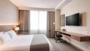 Modern hotel bedroom with neatly made bed, work desk with chair, flat-screen television mounted on wall, neutral beige and gray color scheme, natural window light, professional business-traveler environment