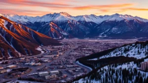 Aerial view of Ouray Colorado nestled in San Juan Mountains with snow-capped peaks surrounding small alpine town during golden hour sunset, dramatic mountain landscape, residential and hotel buildings visible below