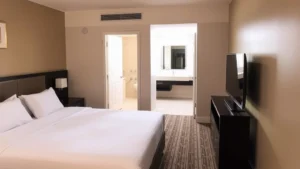 Modern hotel room interior with queen bed, flat-screen TV mounted on wall, neutral beige tones, clean contemporary bathroom visible through doorway, natural morning light from window, no visible text or signage