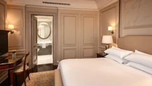 Luxurious hotel room with king bed, marble bathroom visible through open door, elegant European décor, soft ambient lighting, high thread count white linens