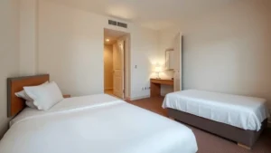 Clean, modest hotel room with two beds, white linens, simple nightstand with lamp, neutral walls, small desk area, and open door showing hallway - photorealistic budget hotel interior during daytime