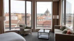 Modern hotel room interior with contemporary furnishings, neutral color palette, floor-to-ceiling windows overlooking urban Roman rooftops and distant church domes, professional photography lighting, no visible text or signage
