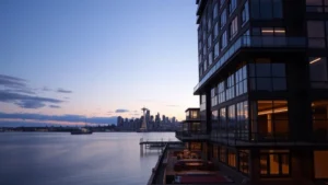 Modern waterfront hotel exterior overlooking Elliott Bay at dusk with Seattle skyline visible across the water, contemporary architecture with glass and steel features, evening lighting reflecting on calm water, Ballard neighborhood location