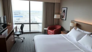 Modern airport hotel room with comfortable bed, work desk, and clean contemporary furnishings overlooking airport grounds, natural daylight through large window, professional business traveler aesthetic, no visible text or signage