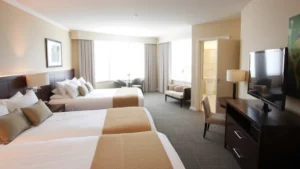 Modern comfortable family hotel suite with two queen beds, contemporary neutral decor, flat-screen TV, and clean bright bathroom visible through open door, natural daylight from windows